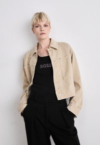 Bežna jeans jakna s kragnatim dizajnom, prednjim džepovima in srebrnimi pritisnimi gumbi, nosi se čez črn top s tekstom iz bleščic. Črne krojene hlače.