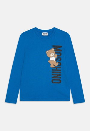 Camisa de manga larga azul para niños con un dibujo de un osito de peluche marrón y texto vertical negro "MOSCHINO" en la parte delantera.