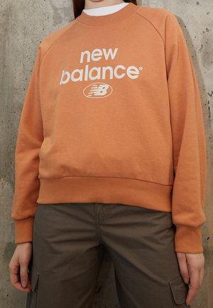 Orange sweatshirt med vitt "new balance"-logotyp och rund emblem. Har ribbade ärmslut och nederkant, avslappnad passform och mjuk tygkvalitet.