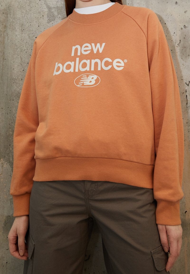 Felpa arancione con logo bianco "new balance" e emblema rotondo. Presenta polsini e orlo a costine, vestibilità rilassata e texture del tessuto morbida.