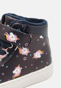 Zapatillas de deporte de cuero sintético negro con un interior de terciopelo azul marino. Presenta un patrón de unicornios y estrellas, suela blanca y una tira de Velcro para el cierre.