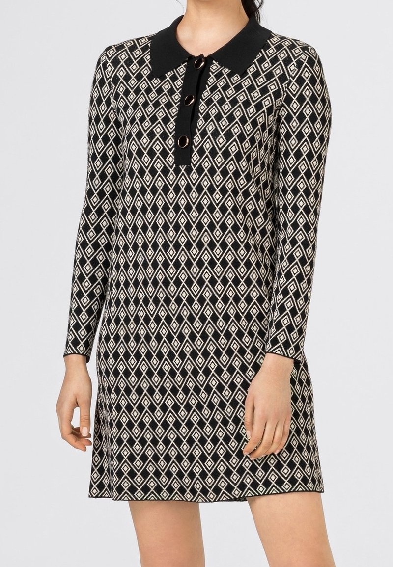 Robe à manches longues en noir et blanc avec motif géométrique en losanges, col noir, et trois grands boutons sur le haut de la poitrine.