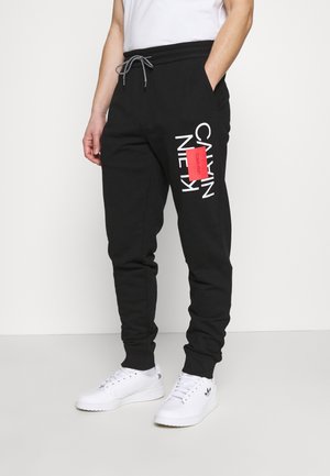 Muž v černých teplácích Calvin Klein s nastavitelným pasem a bílými teniskami, stojící před bílým pozadím.