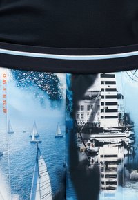 Μπλε ρύθμιση ρούχου πάνω από ύφασμα με εκτύπωση μπλε θάλασσας και ιστιοφόρων στα αριστερά, φάρος με μαύρες και λευκές ρίγες και κτήριο στα δεξιά.