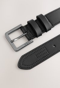 Melna ādas josta ar taisnstūrveida sudraba sprādzi, uz kuras iegravēts "NEXT", ar izšūtiem akcentiem un diviem jostas cilpām uz bēša fona.