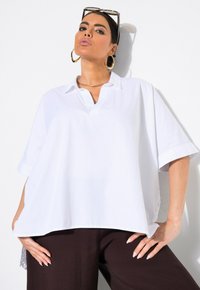 Camicia bianca di cotone oversize con colletto ribattuto, maniche corte e spacco laterale. Abbinata a pantaloni marroni scuri con tessuto testurizzato.