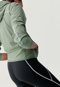 Svetlozelená atletická bunda s reflexnými akcentmi, v kombinácii s čiernymi legínami s bielymi zakrivenými líniami; hladká textúra a priliehavý strih.