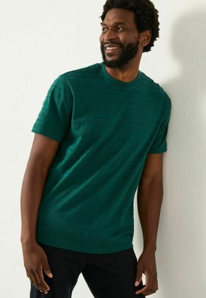 Grünes Kurzarm-T-Shirt mit strukturiertem Muster, Rundhalsausschnitt und entspannter Passform. Getragen mit dunklen Hosen, was einen lässigen Stil zeigt.