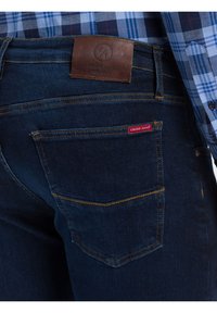 Tmavě modré džíny s oranžovým prošitím, koženou nášivkou značky na pasu a červenou etiketou na zadní kapse, doplněné modrou károvanou košilí.