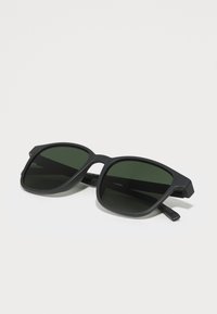 Ochelari de soare negri cu un finisaj mat, lentile pătrate și sticlă cu nuanță verde. Au brațe groase cu o textură subtilă pentru aderență.