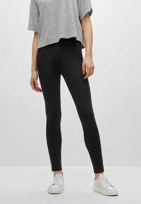 Leggings noirs en matière extensible, avec une coupe ajustée et des zips aux chevilles, associés à un T-shirt court gris et des baskets blanches.