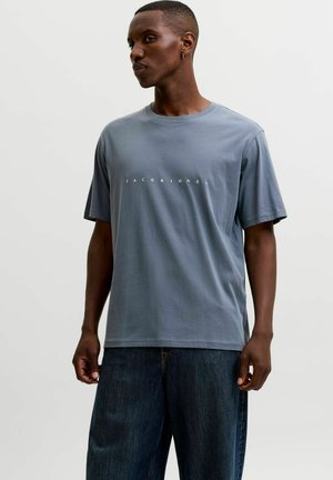 Homme noir portant un t-shirt bleu Jack & Jones et un jean bleu foncé, debout devant un fond gris clair uni.