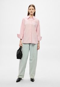 Camisa de botões às riscas rosas e brancas com mangas largas, combinada com calças largas verde claro e uma bolsa preta. Sapatos pretos polidos.