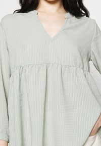 Blouse vert clair avec un décolleté en V, des manches longues, un tissu texturé et une taille froncée. Présente des rayures verticales subtiles et des poches latérales.