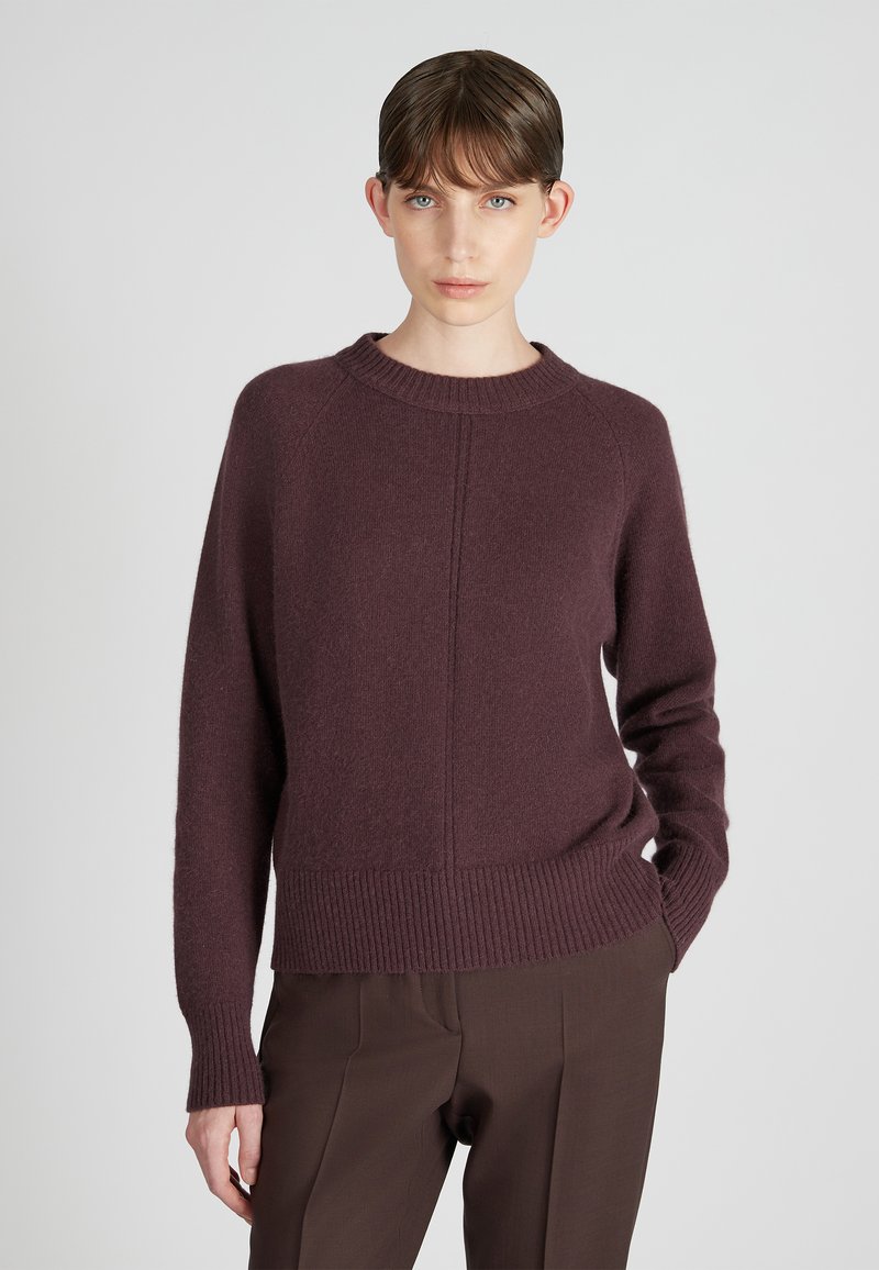 Maglione in lana bordeaux con colletto a girocollo, polsini e orlo a coste, caratterizzato da una texture liscia e un sottile dettaglio di cucitura frontale. Indossato con pantaloni marroni.