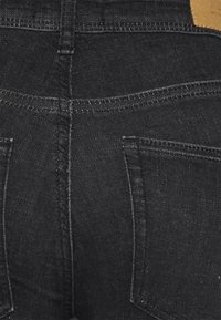 Jeans en denim noir avec une silhouette ajustée, des coutures distinctives et deux poches arrière. Le matériau a une texture lisse.
