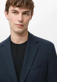 Jonge man met kort bruin haar, draagt een donkerblauwe blazer over een zwart shirt met ronde hals, kijkt iets naar de zijkant tegen een witte achtergrond.