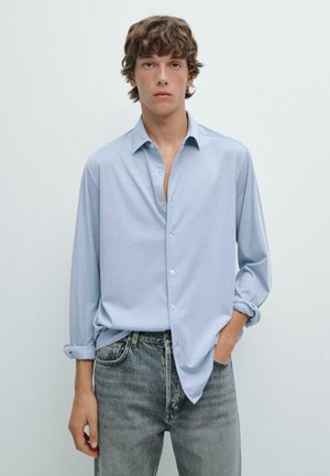 Light blue button-up shirt in a soft, smooth fabric; features a classic collar and rolled sleeves, paired with gray, distressed jeans.
