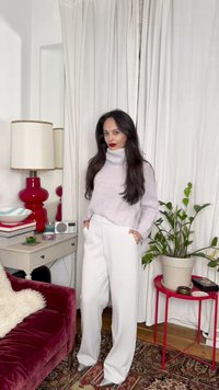 Moteris su ilgais tamsiais plaukais dėvi baltą golfą ir plačias baltas kelnes, stovinti svetainėje su raudonu aksominiu krėsliu ir žalia augalu.