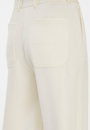 Nahaufnahme von cremefarbenen Hosen, die hinten Taschen mit dezenten Nähten und Gürtelschlaufen vor einem einfarbigen Hintergrund zeigen.