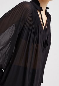 Blusa nera trasparente con dettagli plissettati, colletto alto e maniche lunghe e vaporose. Presenta un nastro al collo e una texture fine.