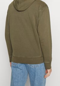 Sudadera con capucha de color verde oliva con dobladillo y puños acanalados, con capucha con cordón y textura suave, combinada con vaqueros azul claro.