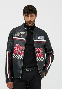 Giacca da corsa in pelle nera con strisce rosse e bianche, motivo a scacchi, patch con logo e chiusura con zip.
