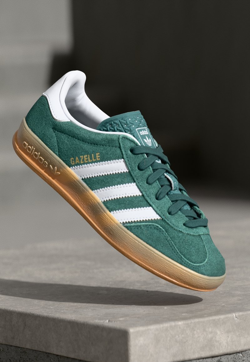 Zelená teniska Adidas Gazelle s bílými pruhy a gumovou podrážkou, zobrazená zavěšená nad betonovým povrchem na tmavém rozmazaném pozadí.
