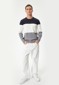 Maglione a righe navy, bianco e grigio con scollo a girocollo, abbinato a jeans bianchi e sneakers bianche, in piedi su uno sfondo neutro.