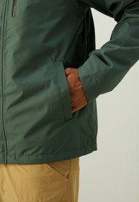 Chaqueta impermeable verde con una textura suave, que presenta un bolsillo con cremallera y un dobladillo ajustable. Combinada con pantalones beiges.