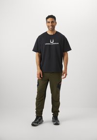 T-shirt noir à manches courtes avec le logo "UNDER ARMOUR", accompagné d'un pantalon de sport vert olive avec des accents noirs et des détails rouges.