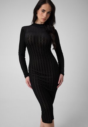 Femme aux longs cheveux foncés portant une robe noire ajustée à manches longues et à motifs, debout devant un fond gris uni.