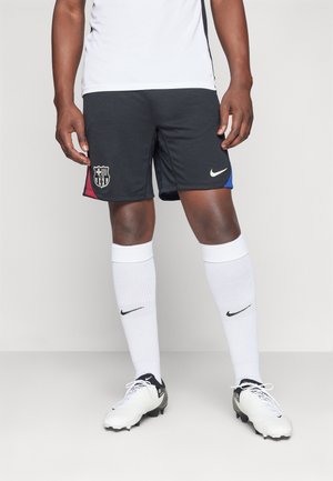 Athlète portant un short noir du FC Barcelone avec les logos du club et de Nike, des chaussettes blanches Nike et des chaussures à crampons blanches sur un fond uni.
