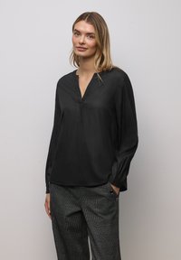 Top nero a maniche lunghe con scollo a V e leggero effetto lucido; presenta una rifinitura decorativa lungo il collo. Indossato con pantaloni grigi a motivo.