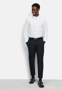 Weißes Hemd mit Knöpfen in Schwarz, kombiniert mit dunklen Hosen und schwarzen Schuhen. Glatter Stoff, taillierter Schnitt und minimalistische Designelemente.