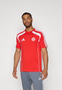 Red athletic shirt made of smooth fabric, featuring white accents and the FC Bayern Munich logo on the left chest. Black stripes on shoulders.