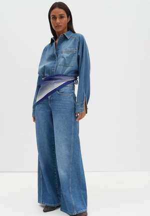 Camicia di denim con chiusura frontale a bottoni e taschino sul petto, abbinata a jeans denim a gamba larga, con cintura fantasia blu e bianca.