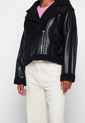 Czarna skórzana kurtka z kożuszkiem, asymetrycznym przednim zamkiem, noszona na różowym topie i białych spodniach.