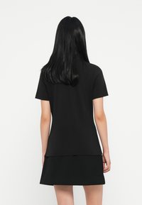 Kvinde med langt sort hår iført en ensfarvet sort kortærmet top og matchende sort nederdel, stående mod en hvid baggrund, med ryggen mod beskueren.