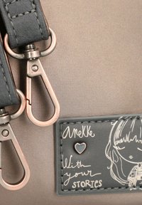 Graue Tasche mit glatter Oberfläche, ausgestattet mit silbernen Beschlägen und zwei Clips. Ein schwarzes Patch zeigt eine weiße Illustration und Text.