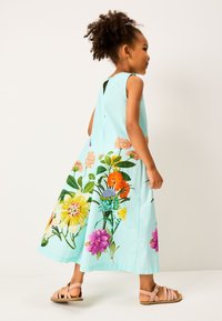 Lichtblauwe mouwloze jurk met levendige bloemenpatronen in oranje, geel, paars en groen. Platte sandalen maken de look compleet.