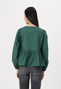 Top peplum verde em algodão com mangas longas bufantes, decote redondo e detalhe de cintura franzida. Usado sobre jeans escuros.
