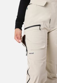 Beige skibukser med sorte lynlåse, udstyret med en sidelomme og RECCO-logo, fremstillet af slidstærkt, vejrbestandigt materiale.