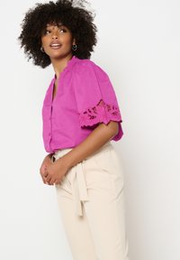 Blusa di lino rosa con maniche a sbuffo e rifiniture in pizzo. Indossata con pantaloni beige a vita alta con cintura legata. Bottoni semplici sul davanti.
