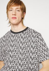 Camiseta gráfica en blanco y negro con el logotipo de "Levi's" impreso repetidamente, mangas cortas y un cuello redondo de canalé negro.