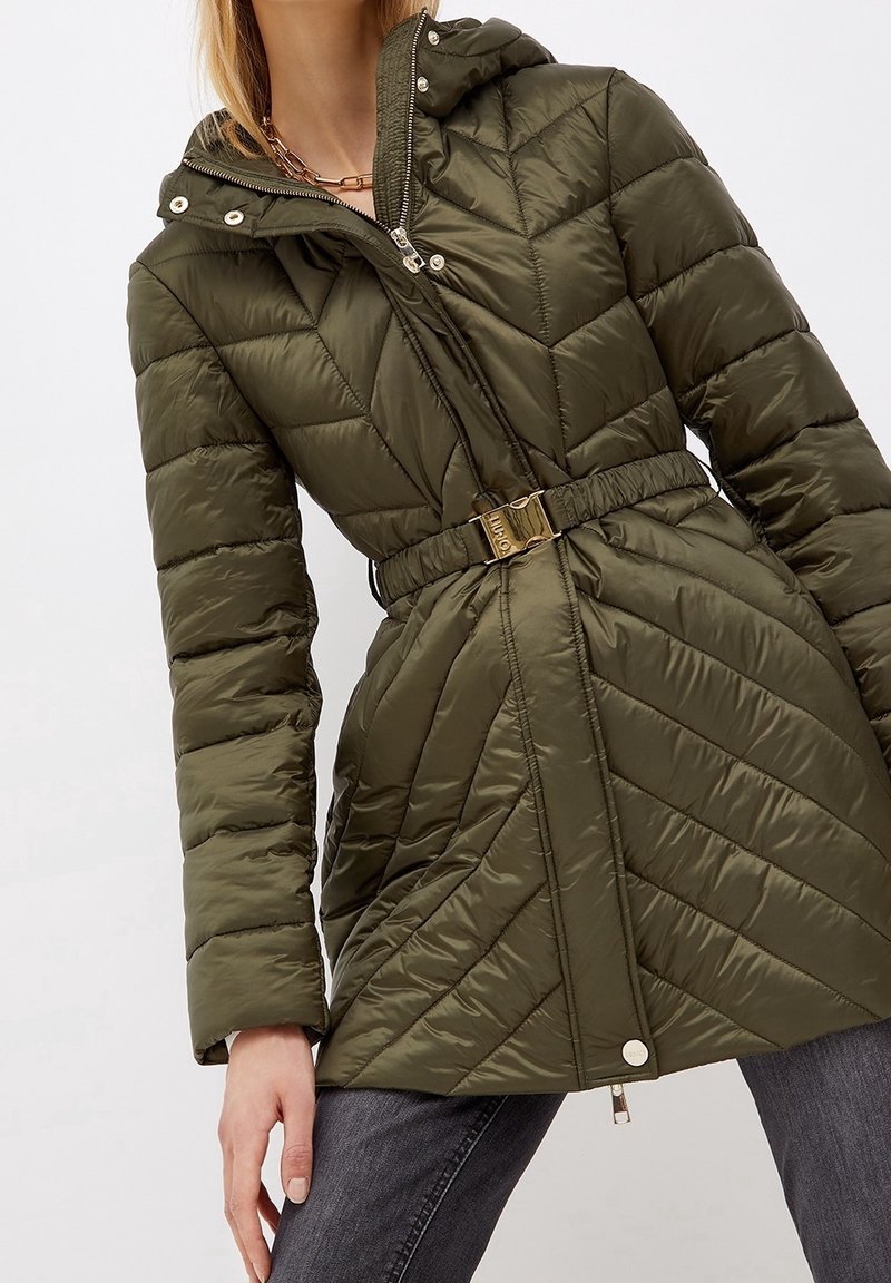 Veste matelassée vert olive avec capuche, taille ceinturée, fermeture éclair métallisée et boutons-pression, portée avec un jean gris foncé.