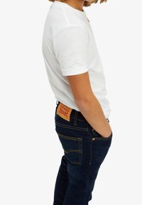 Vit bomulls-t-shirt med korta ärmar och rund hals, kombinerad med mörkblå jeans med gul söm och en ljusbrun läderlapp.