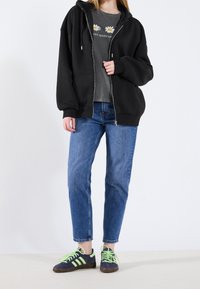 Sweat à capuche noir zippé porté sur un t-shirt gris à imprimé marguerite, associé à un jean bleu clair et des baskets bleu marine avec des lacets verts.