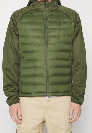 Mann trägt eine olivgrüne, gesteppte Kapuzenjacke mit wattierter Vorderseite und einfarbigen Ärmeln, kombiniert mit beige-farbenen Cargohosen.