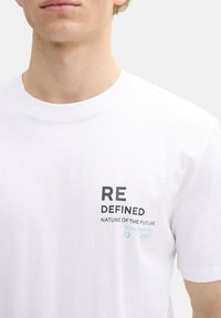 Balta medvilninė marškinėlių su juodu ir žaliu tekstu: "RE DEFINED," "NATURE OF THE FUTURE," ir įsteigimo metai. Klasikinis apvalus iškirpimas.